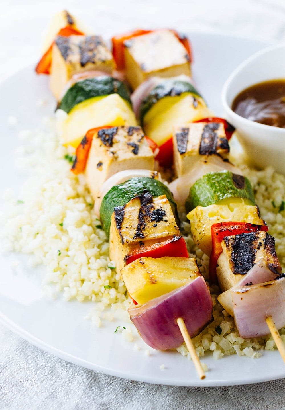 side angle view of grilled tofu skewers on a plate with cauliflower rice and pineapple teriyaki sauce.