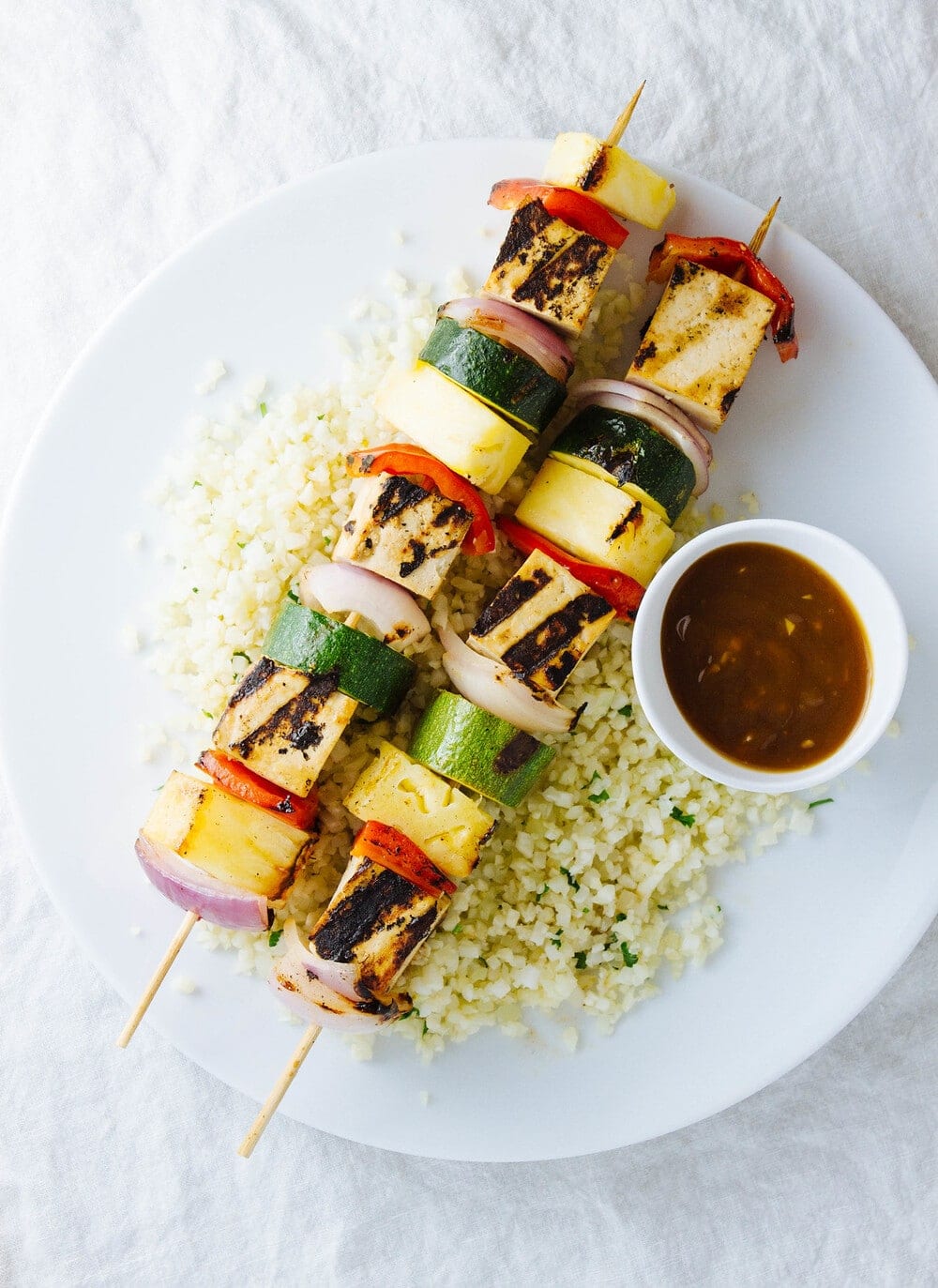 top down view of two grilled tofu skewers on a white plate with side of cauliflower rice and pineapple teriyaki sauce.