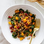 top down view of a serving of autumn wheat berry and butternut squash salad in a white bowl with gold fork.