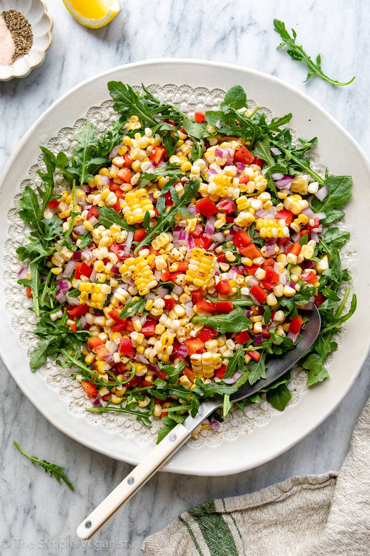 top down view of corn arugula salad on a white serving platter with large spoon and items surrounding.