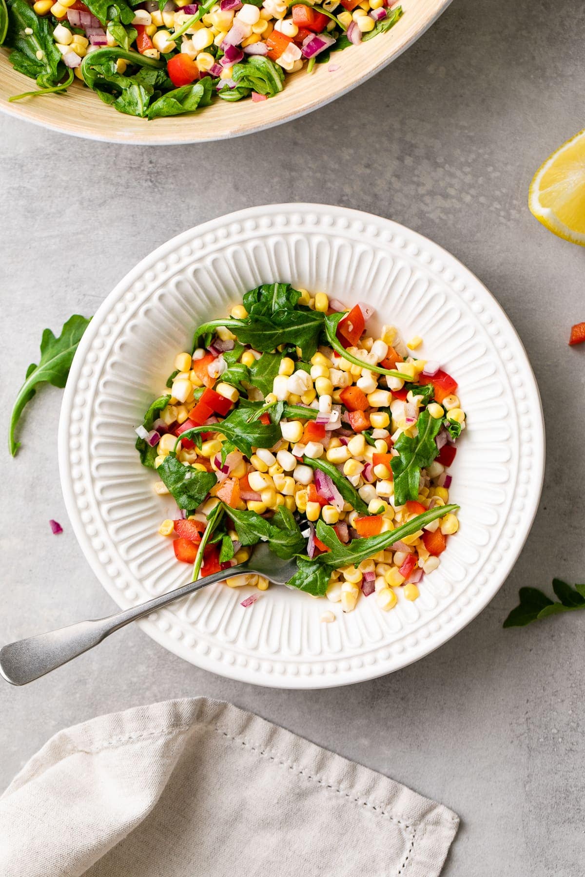 top down view of corn arugula salad in white bowl with fork and items surrounding.
