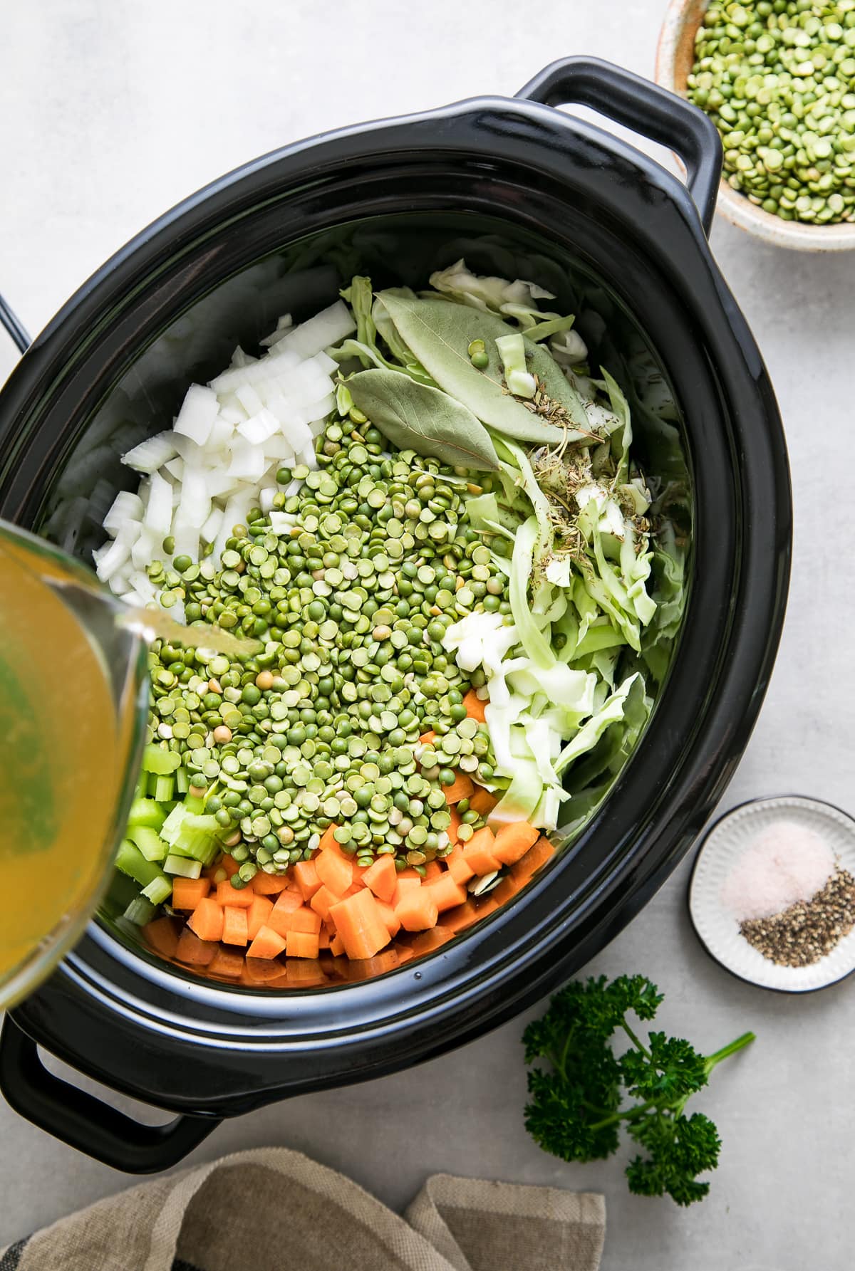 top down view showing the process of adding liquids to pot of split pea soup ingredients in a slow cooker.