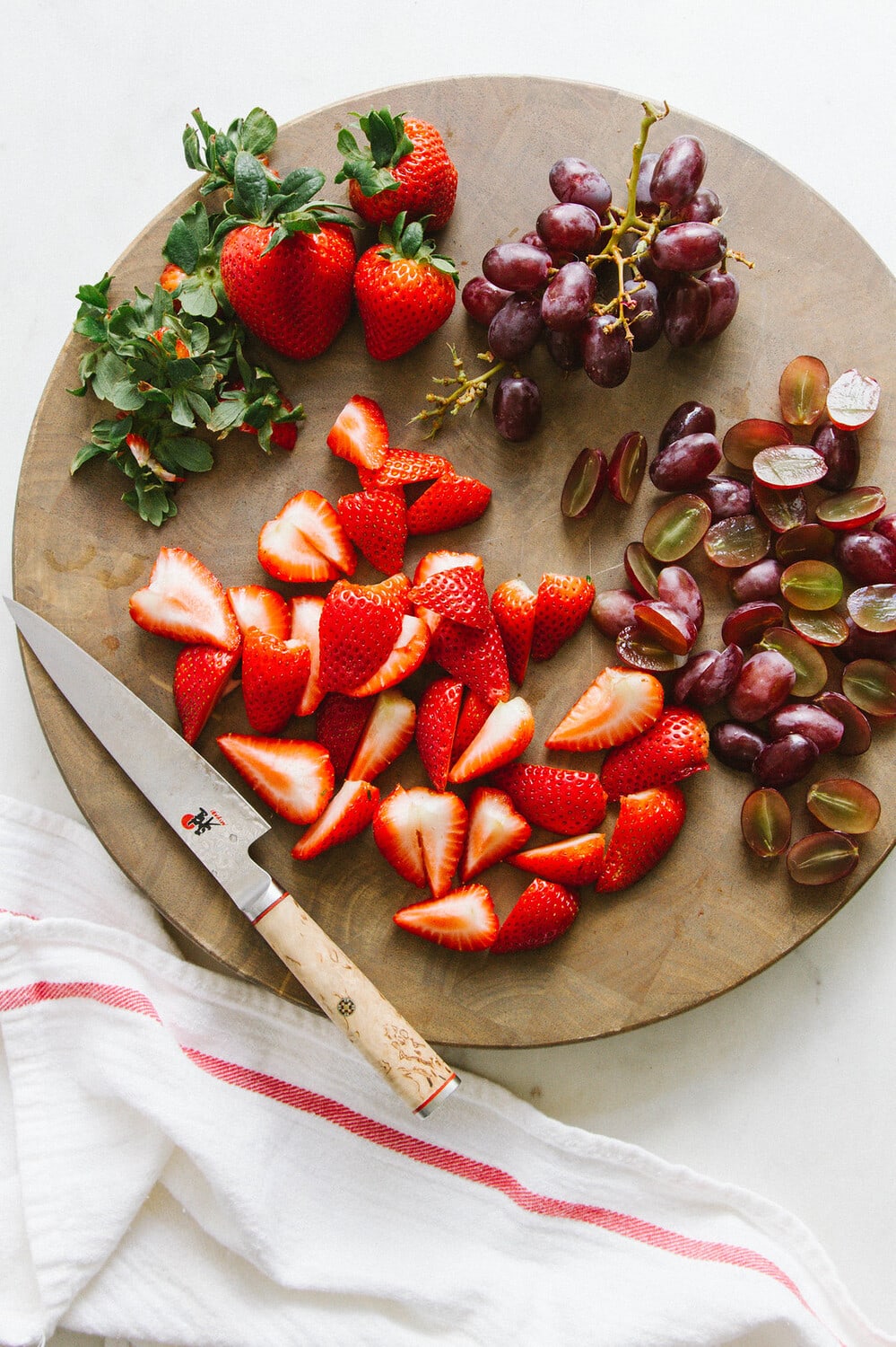 top down view of sliced strawberries and grapes on a circular wooden cutting board.