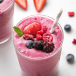 side angle view of healthy mixed berry yogurt smoothie in a glass with straw and items surrounding.