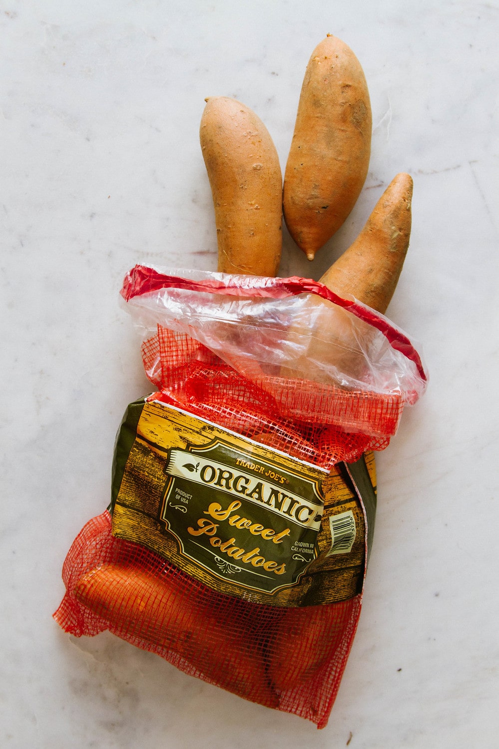 top down view of a bag of sweet potatoes the bag they were purchased in, with a few sticking out of the bag, laying on a marble slab