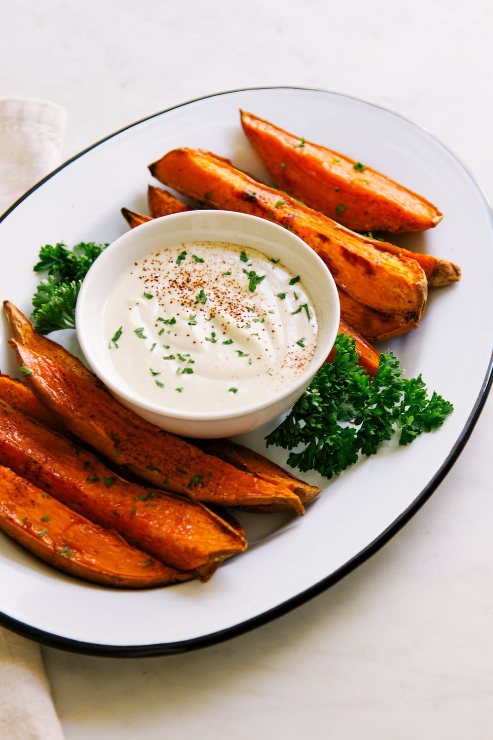 side angle view of baked sweet potato wedges on an oval white plate with black rim with a bowl of vegan aioli in the center