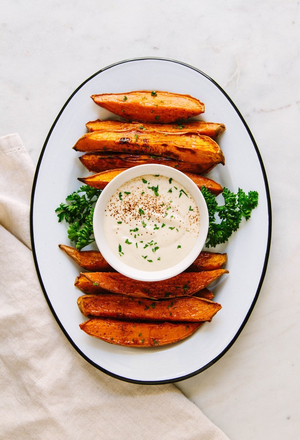 top down view of baked sweet potato wedges and aioli in a small bowl on an oval white plate with black rim