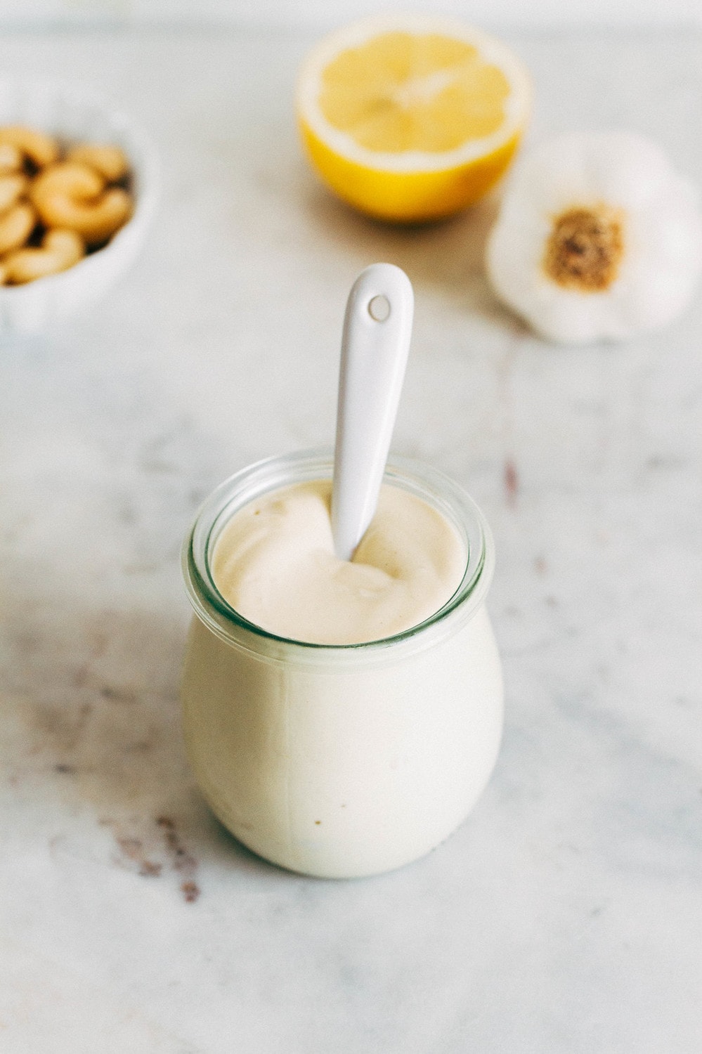 top down angled view of a small glass jar with vegan aioli and spoon, surrounded by a small white bowl of cashews, sliced lemon half and bulb of garlic