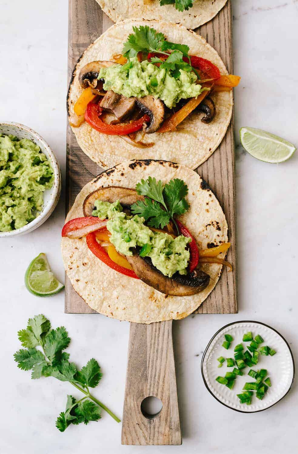 top down view of easy portobello mushroom fajitas topped with jalapeno guacamole on corn tortillas on a wooden serving board.