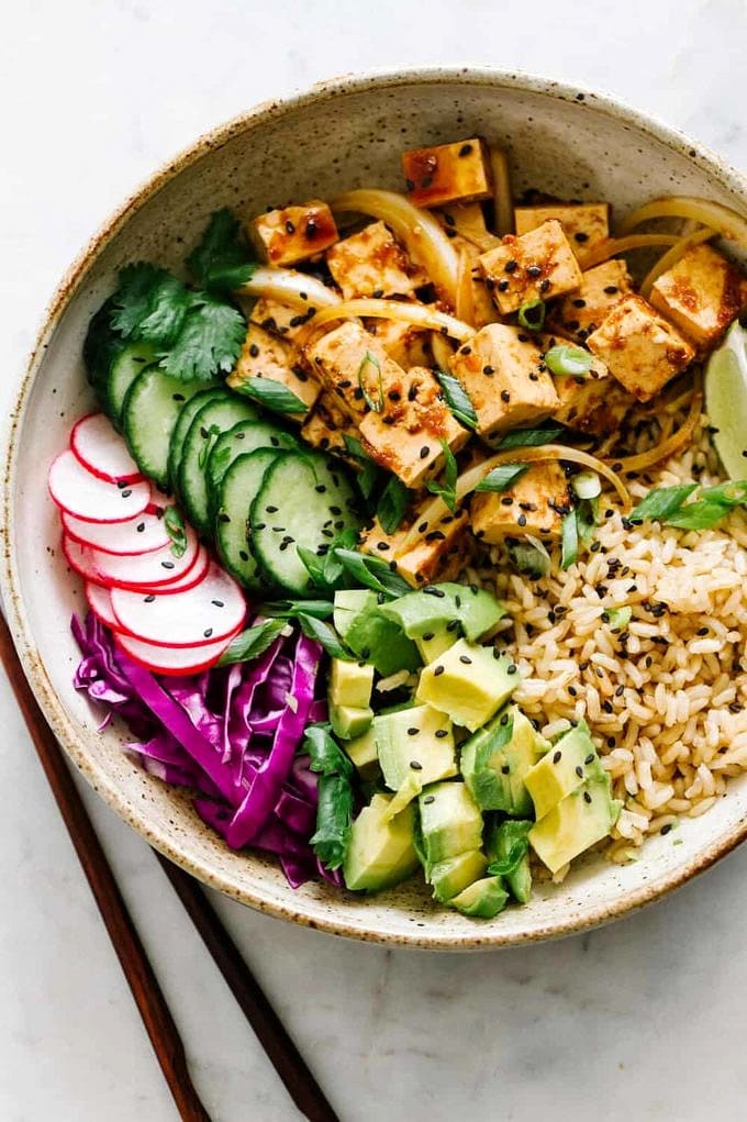 spicy tofu poke bowl with sprouted brown rice, avocado, cucumber, radish, red cabbage, green onions, lime wedges and black sesame seeds