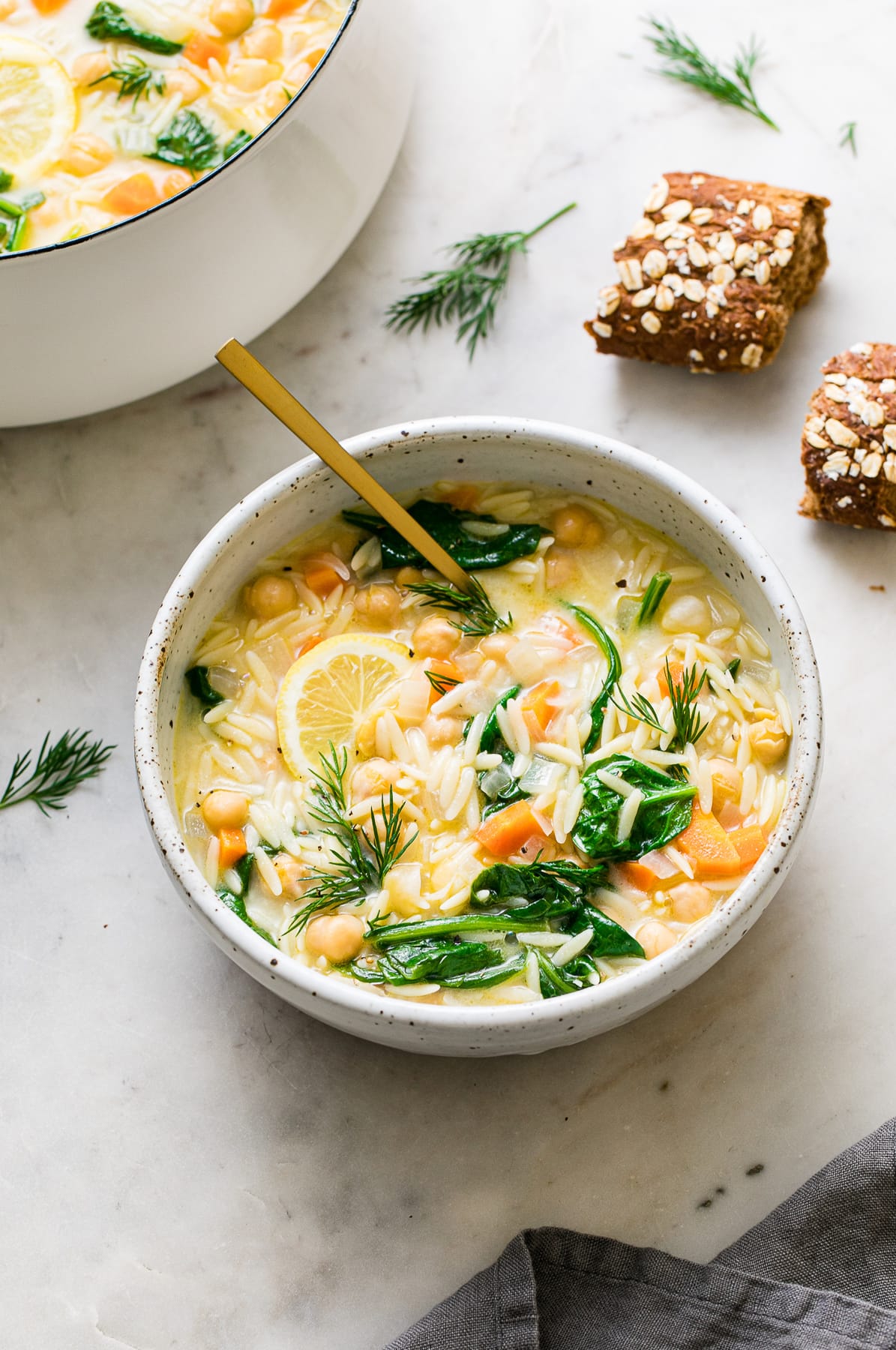 side angle view of bowl full of lemon chickpea orzo soup with gold spoon and items surrounding.