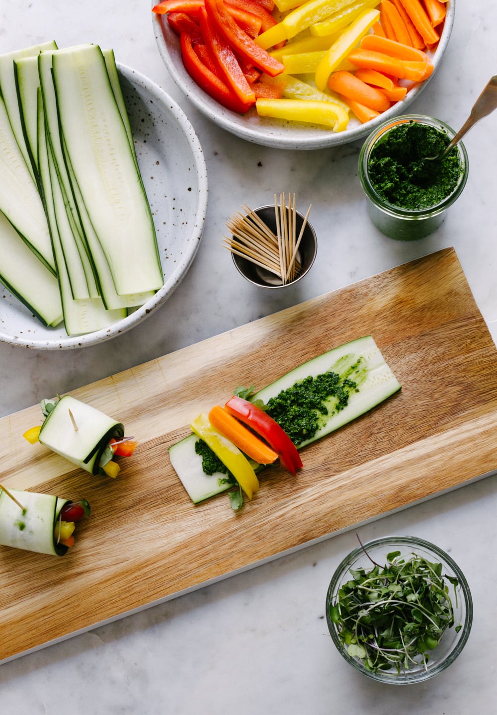 a top down view of making raw zucchini wraps, sliced zucchini, fresh bell peppers, carrots, micro greens and spicy kale pesto, and toothpicks to hold them together once rolled