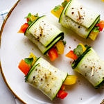 a top down view of raw zucchini wraps filled with fresh bell peppers, carrots, kale pesto and micro greens on a white plate with gold rim