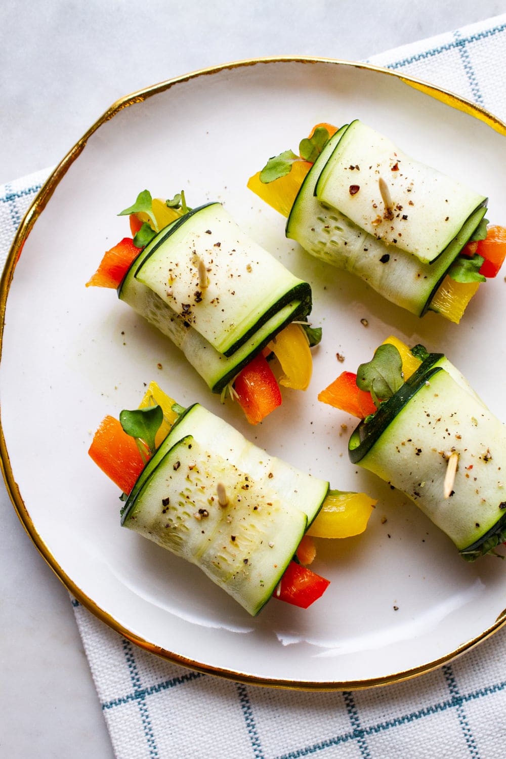 a top down view of raw zucchini wraps filled with fresh bell peppers, carrots, kale pesto and micro greens on a white plate with gold rim