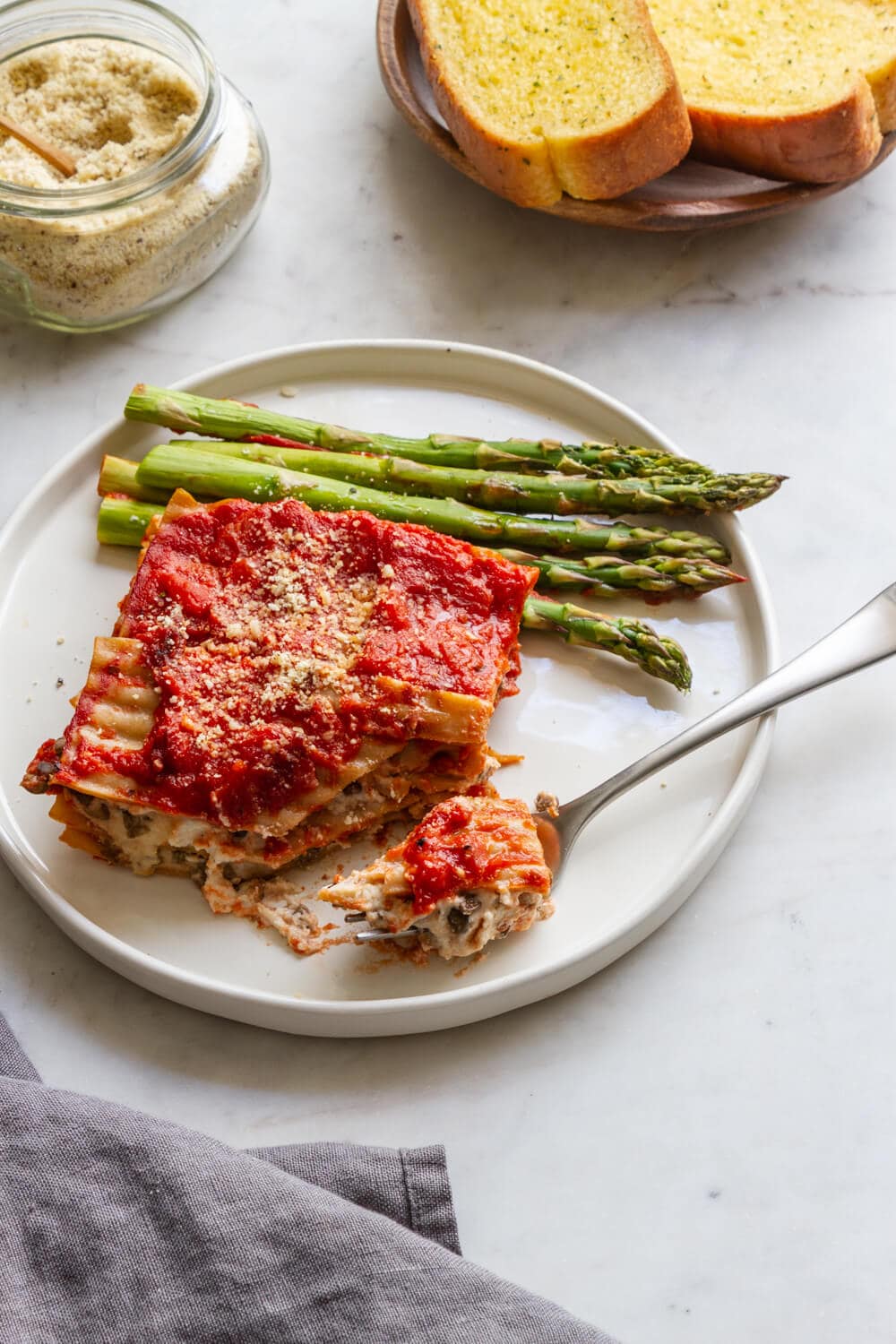 side view of a plate with a slice of simple vegan lasagna and asparagus, with almond parmesan and bread on the side