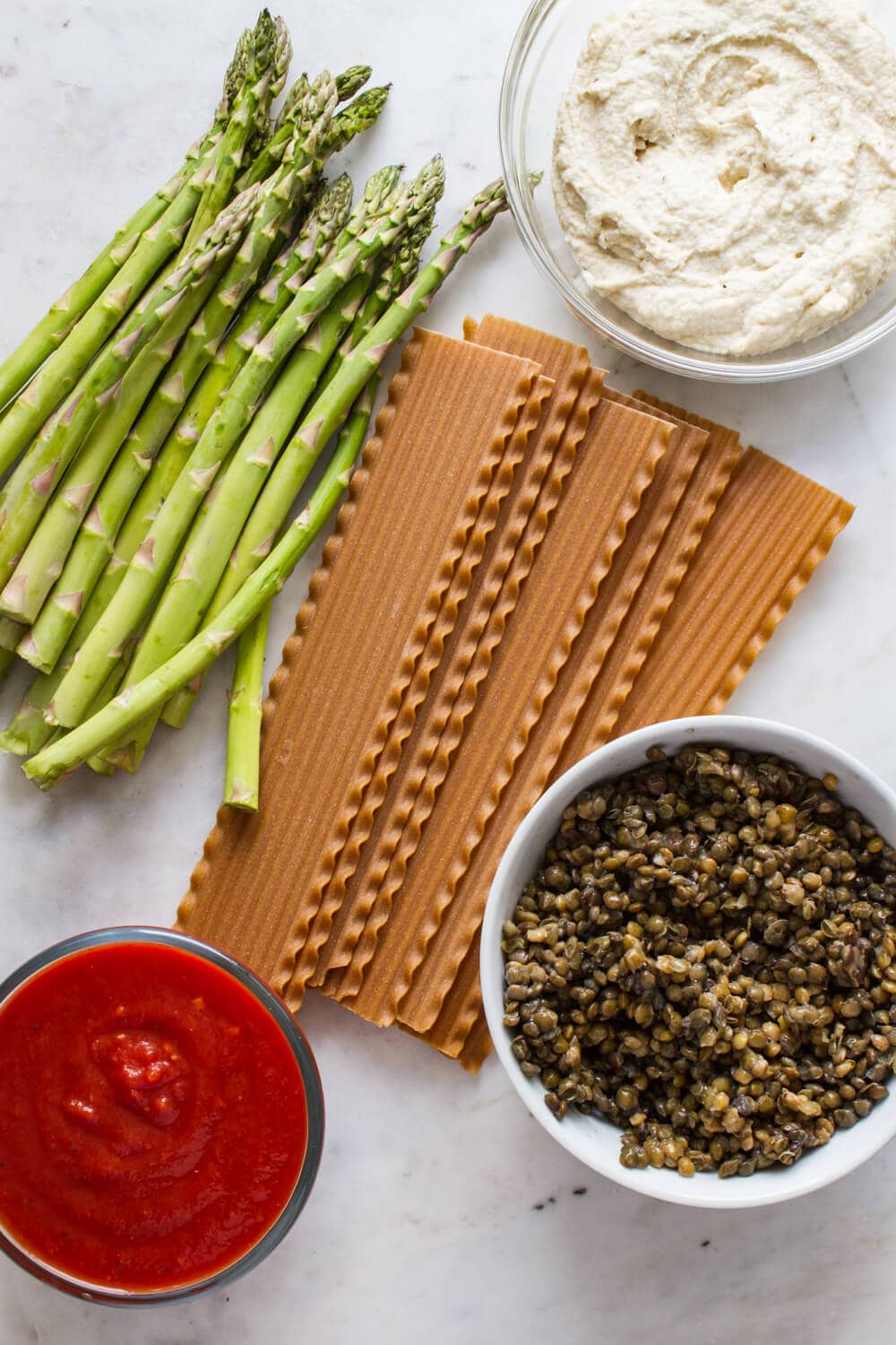 ingredients for vegan lasagna laid on a marble slab, including noodles, pasta sauce, cashew ricotta cheese, lentils and fresh asparagus