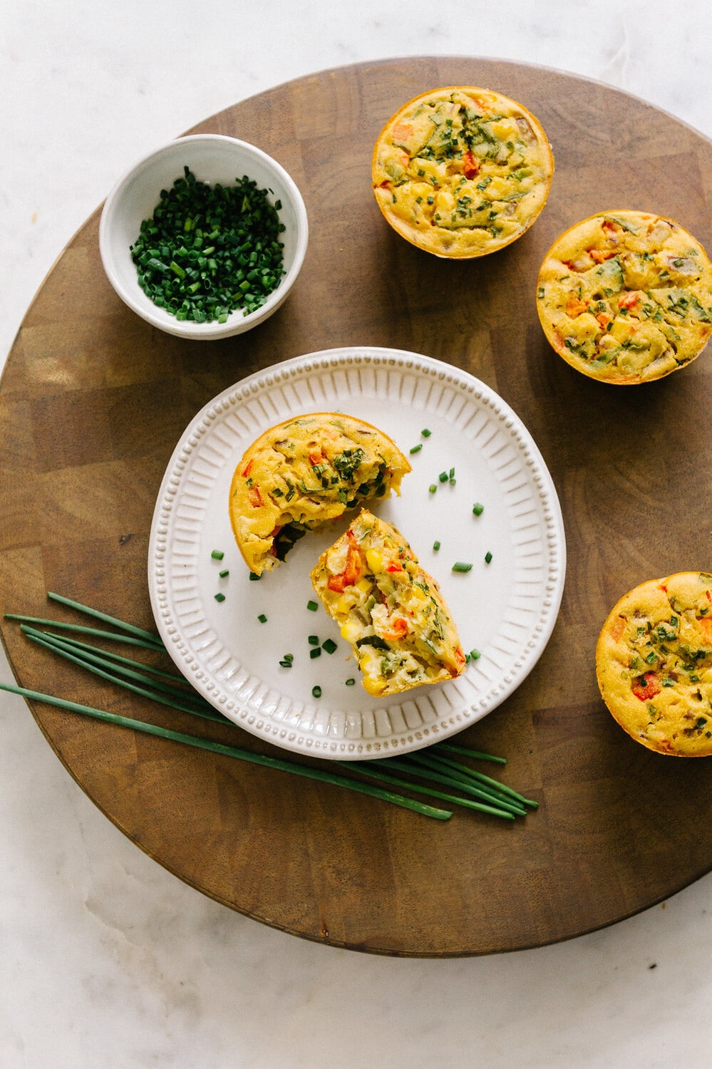 top down view of a mini chickpea flour frittata on a small plate split in half, surrounded by 3 more frittata muffins on a round wooden cutting board