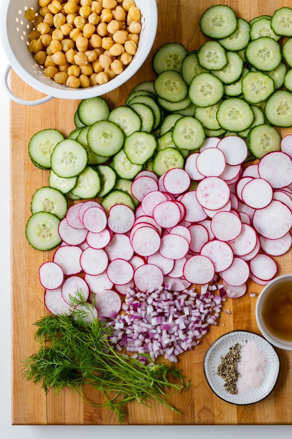 top down view of a wooden cutting board with sliced radishes, cucumbers, diced red onion, whole chickpeas in a small white colander, dill, apple cider vinegar and small dish with salt and pepper