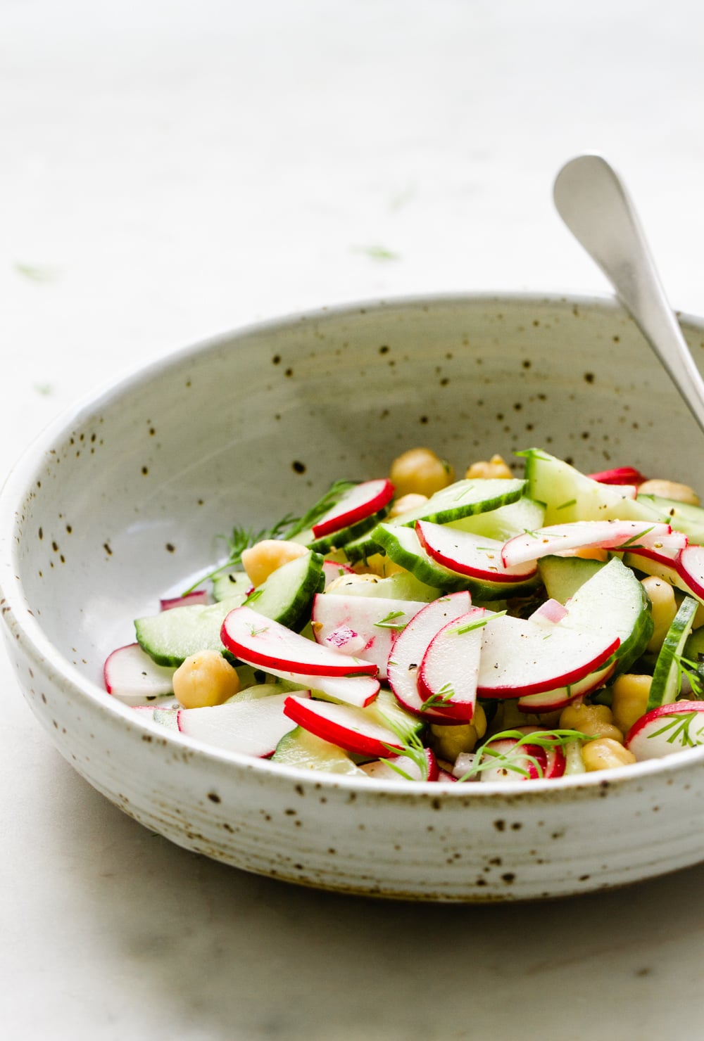 side angel view of a rustic bowl with a serving of radish cucumber salad and spoon on a marble slab