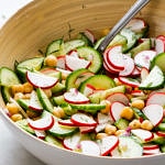 side angle top down view of a white bowl serving bowl filled with radish and cucumber salad with a serving spoon on a marble slab
