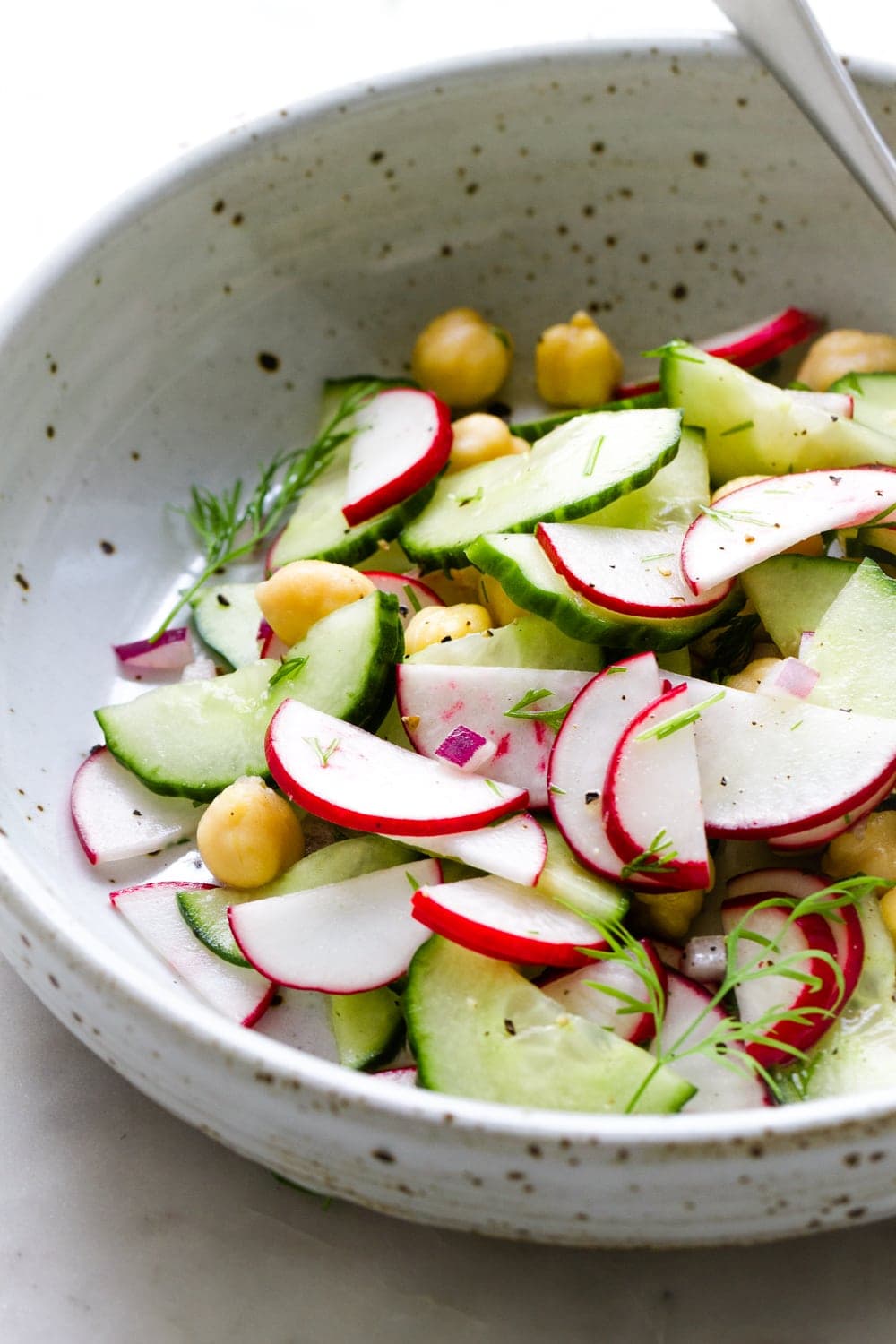 close up, side angle view of a small bowl with a serving of radish cucumber salad and fork on a marble slab