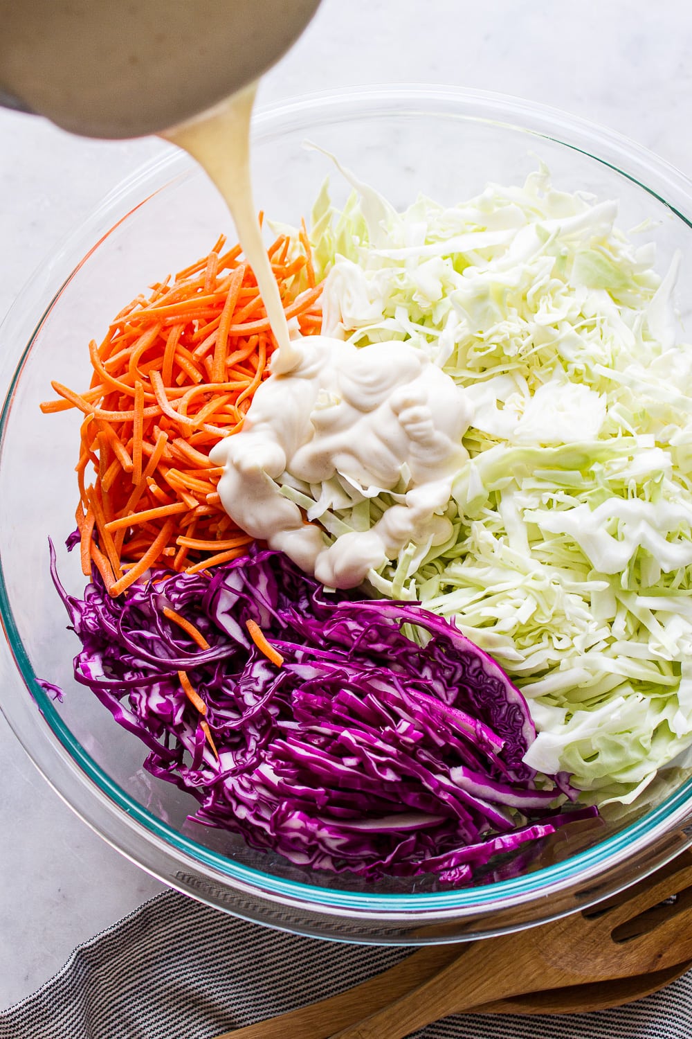 top down view of coleslaw dressing being poured over cabbage and carrots in a large glass mixing bowl.