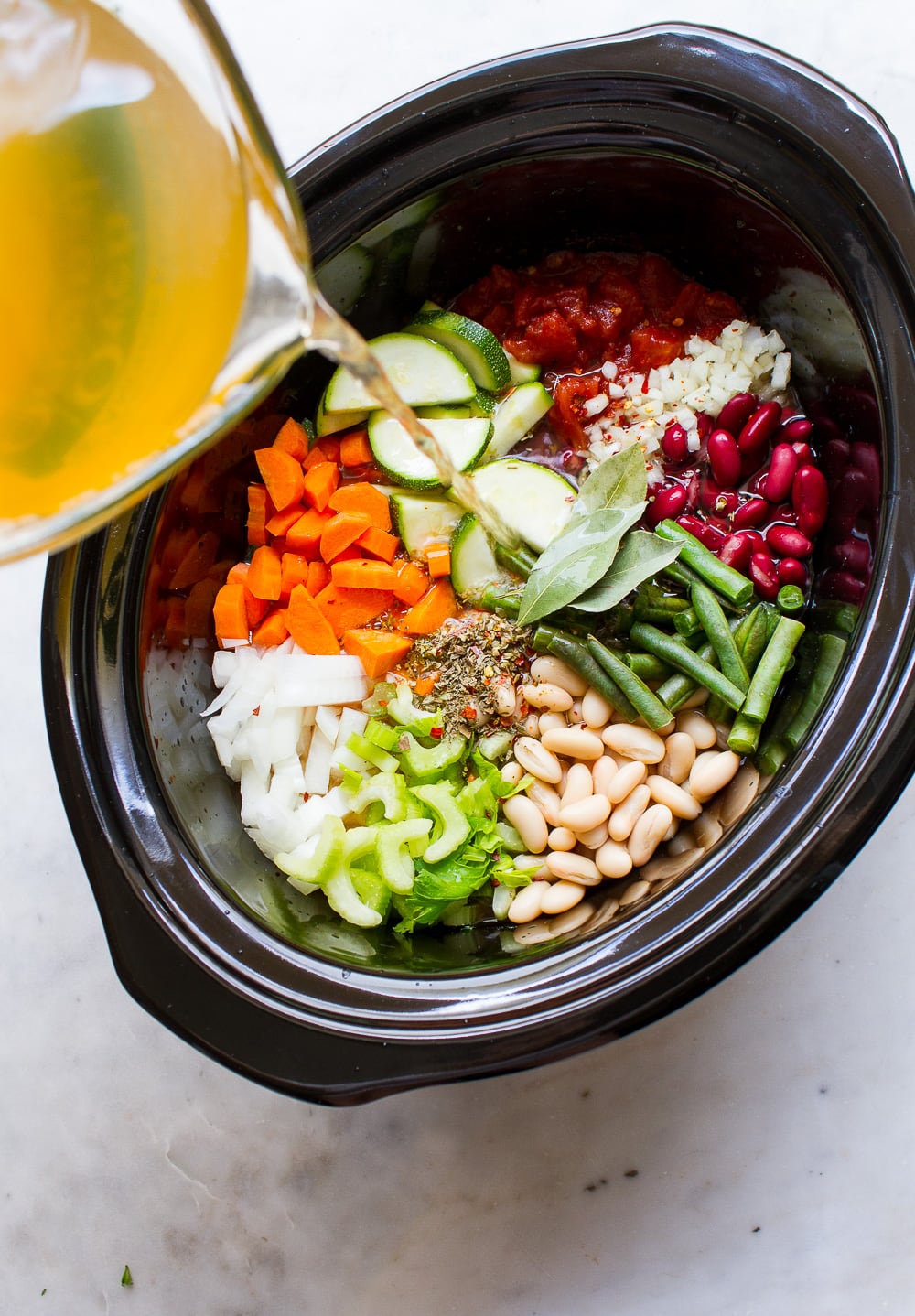 top down view of veggie broth being poured into slow cooker with ingredients for best minestrone soup.