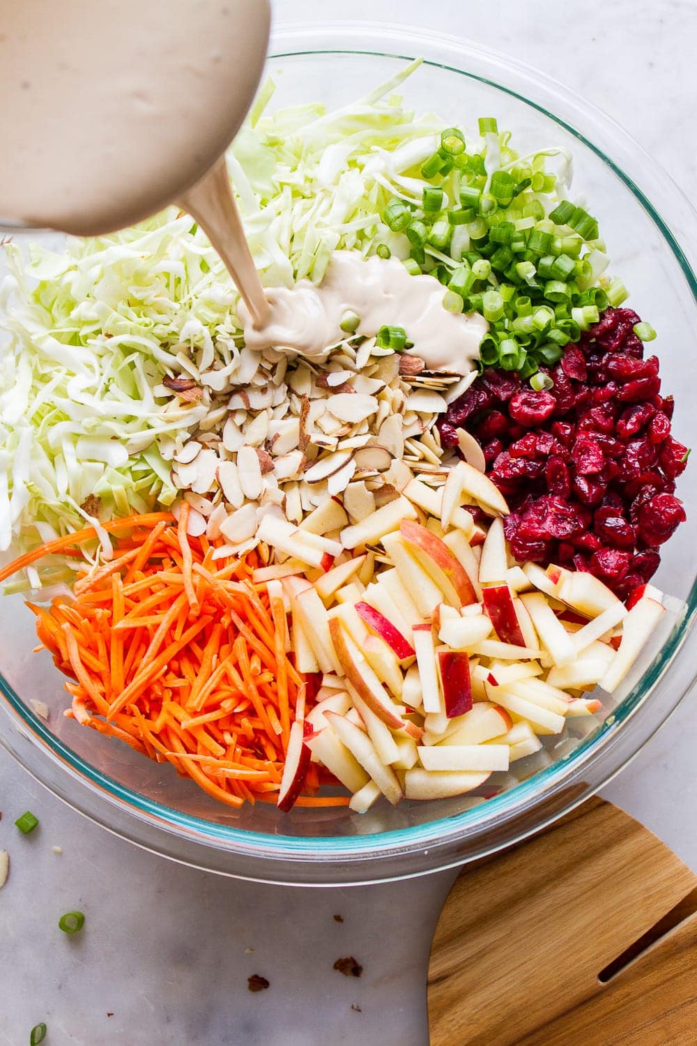 top down view of a large glass mixing with ingredients for apple coleslaw with dressing being poured overtop.