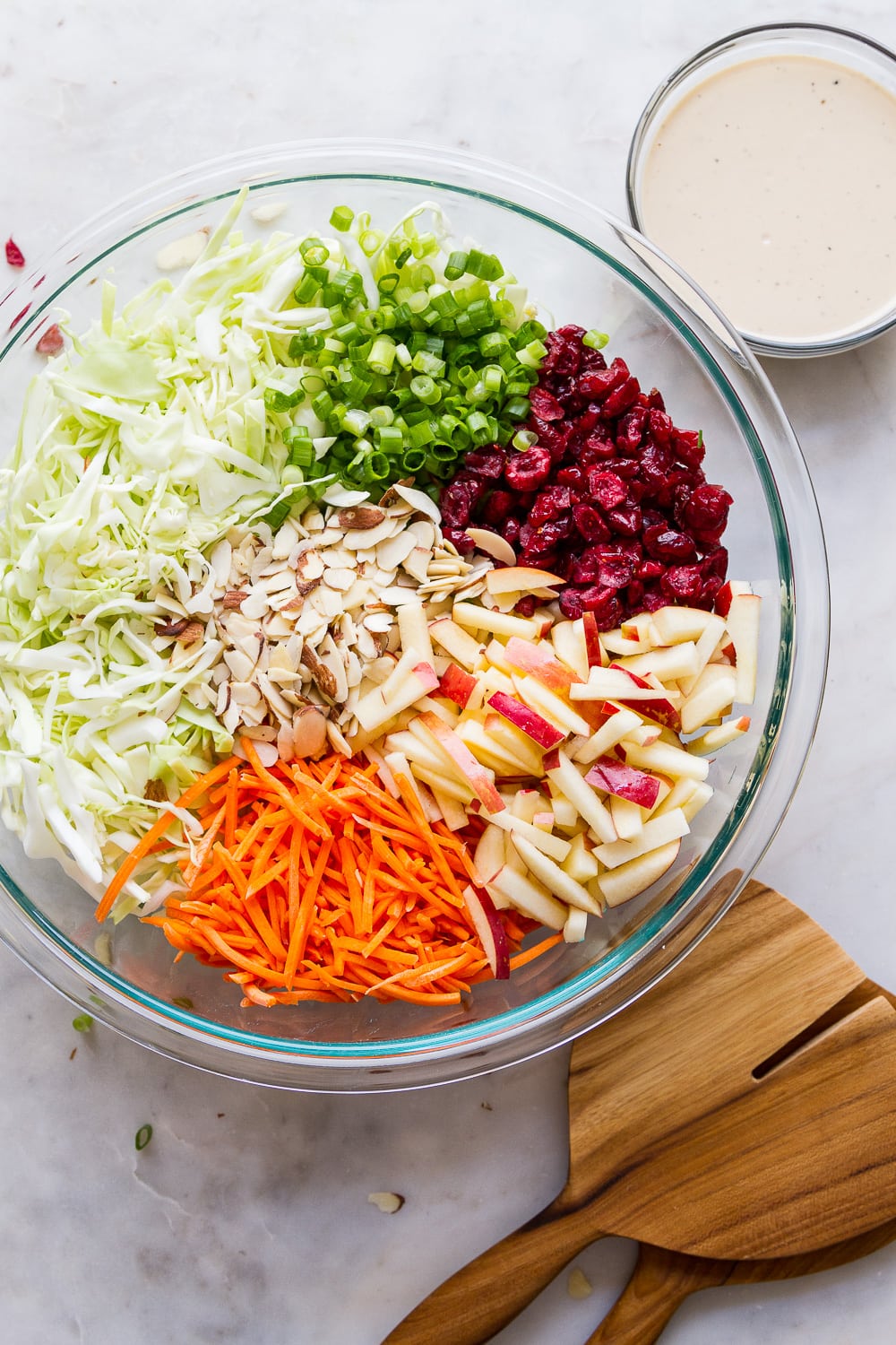 top down view of a large glass mixing with ingredients for apple coleslaw with dressing in a small bowl nearby.
