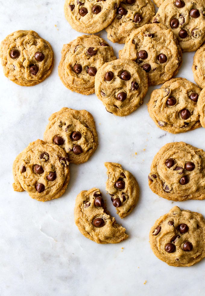 top down view of a vegan chocolate chip cookie broken in half and surrounded by a bunch of vegan chocolate chip cookies on a marble slab.