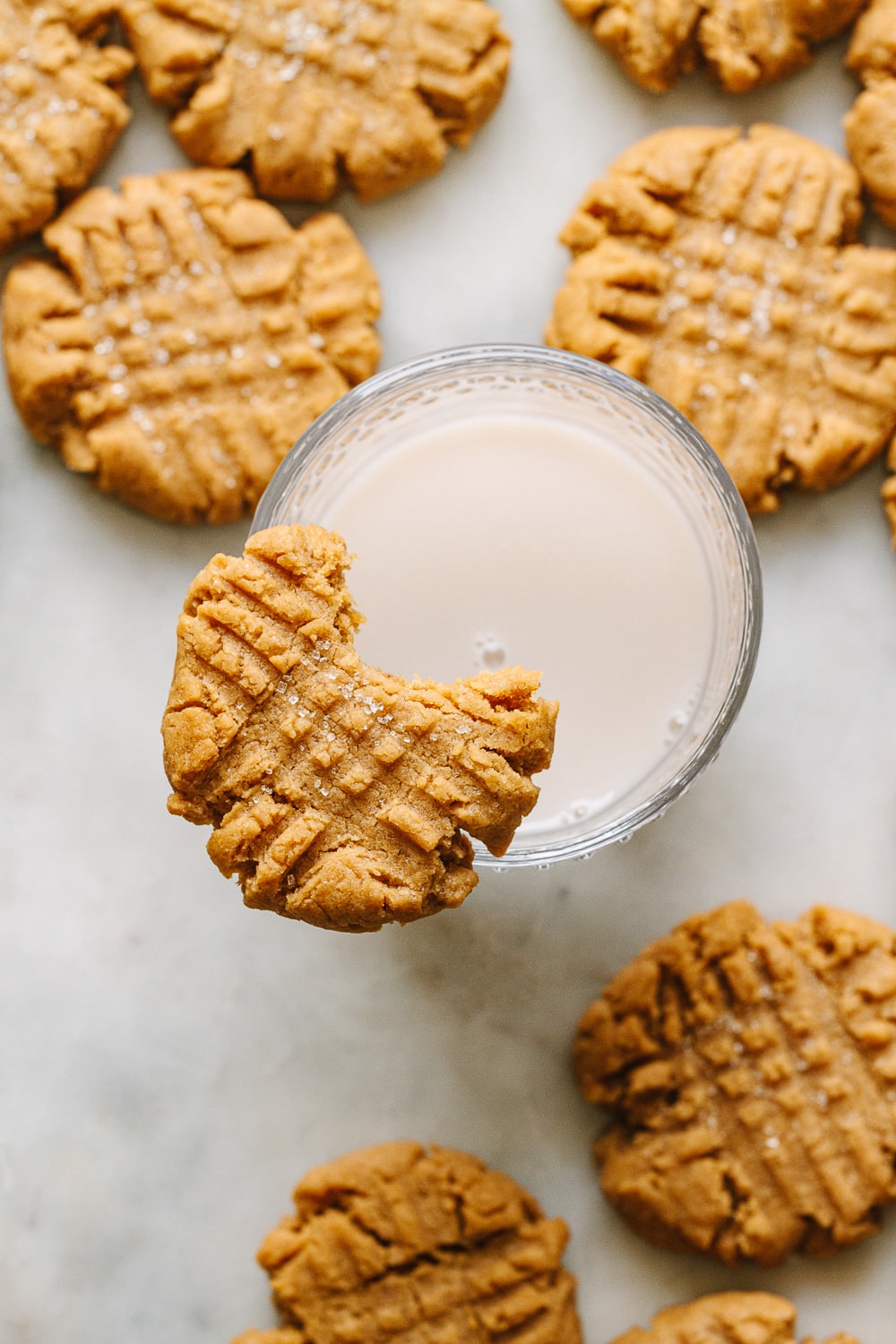 down view of a vegan peanut butter cookie with bite taken out resting on a glass of almond milk with things surrounding.