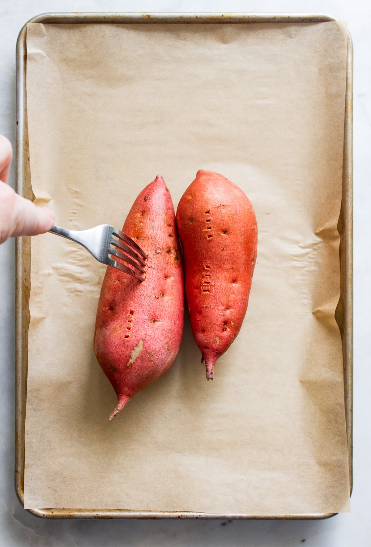 top down view of 2 sweet potatoes on a rimmed baking sheet with holes poked using a fork.