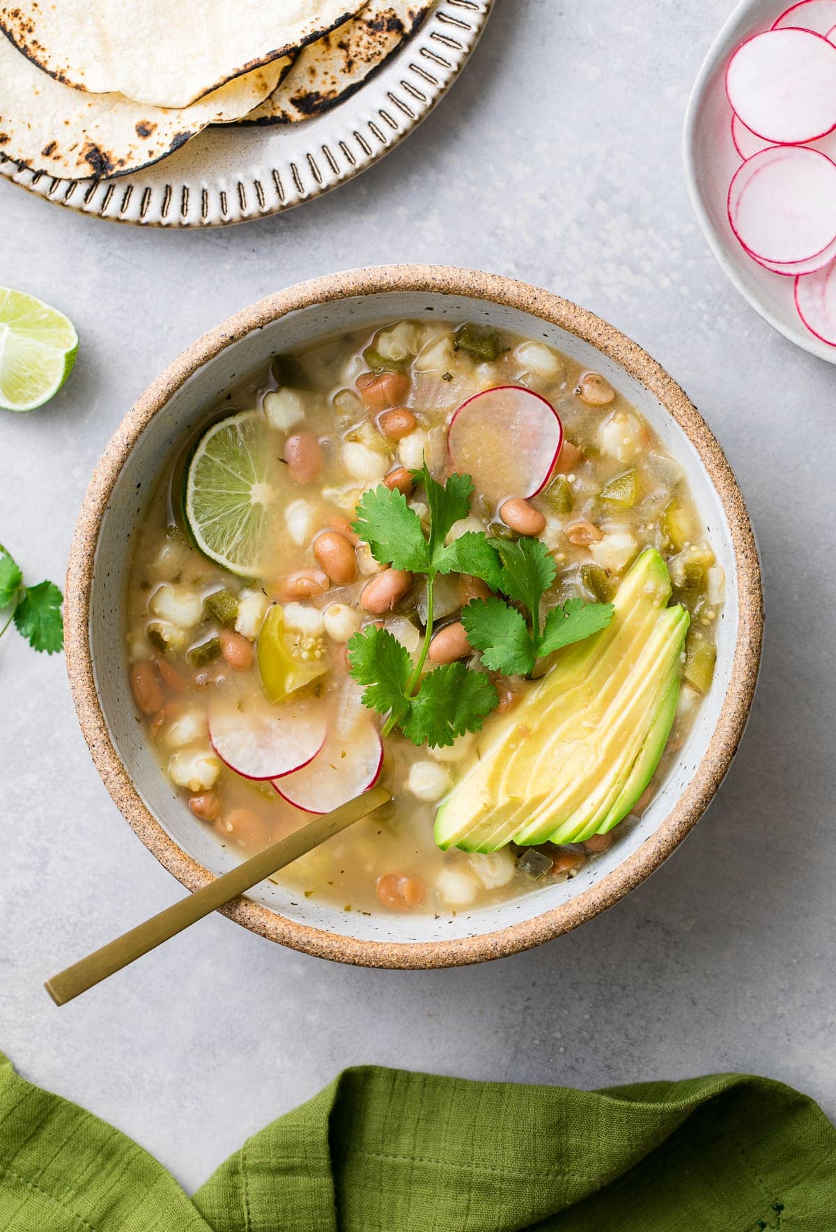 top down view of bowl with posole verde with spoon and items surrounding.