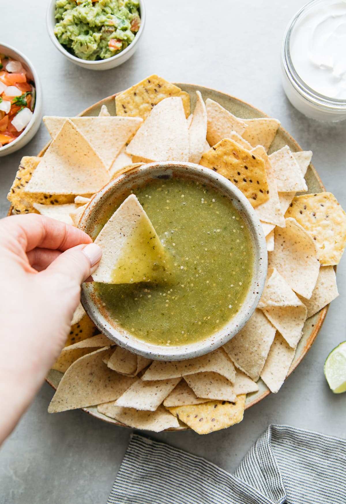 top down view of chip being dipped into bowl filled with tomatillo verde sauce surrounding by tortilla chips and other items.