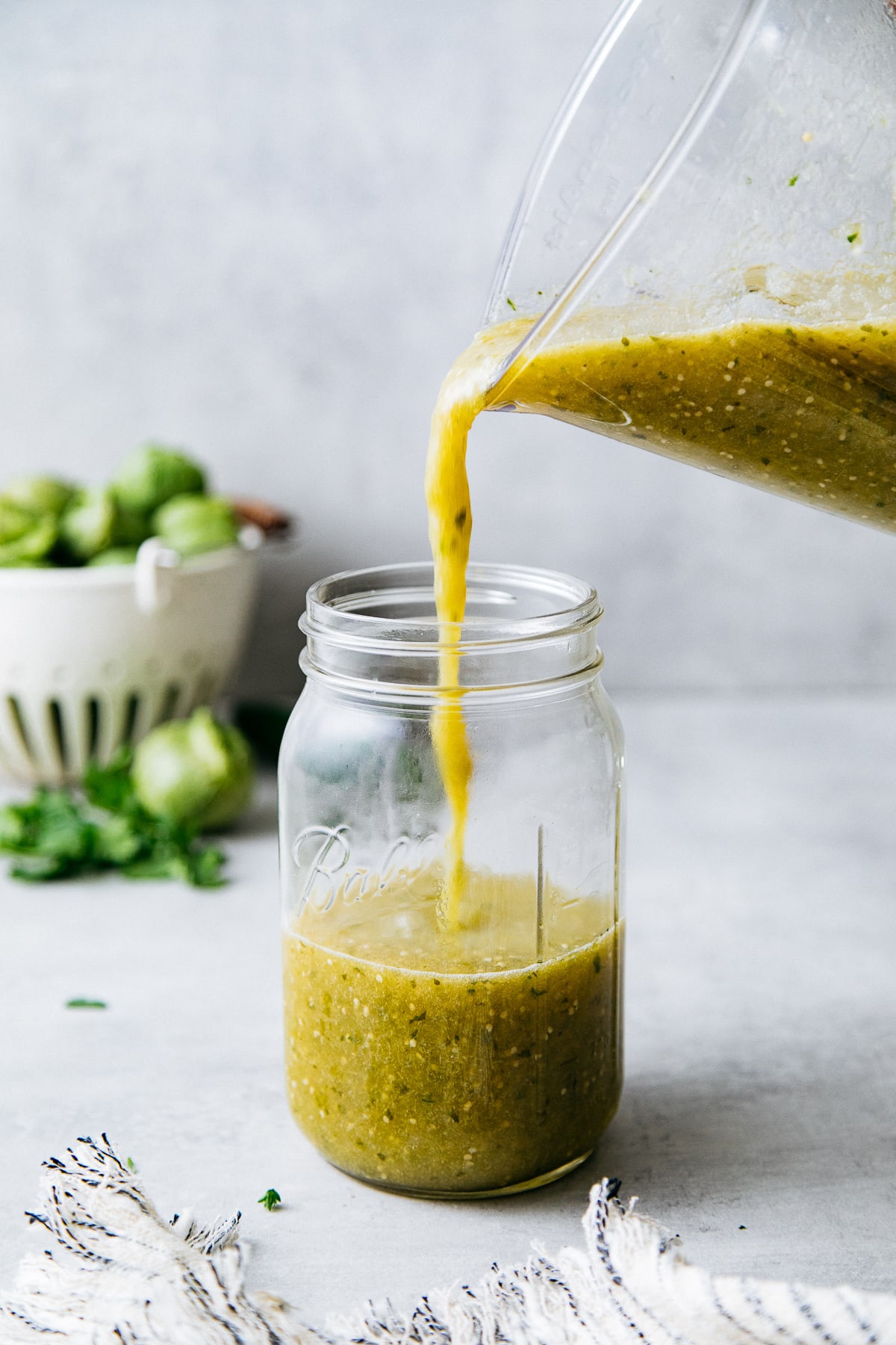 head on view of verde sauce being poured into a glass jar with items in the background.