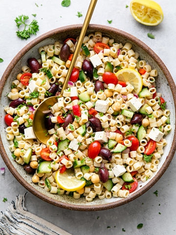 top down view of healthy, vegan Greek pasta salad in a serving bowl with gold serving spoon.