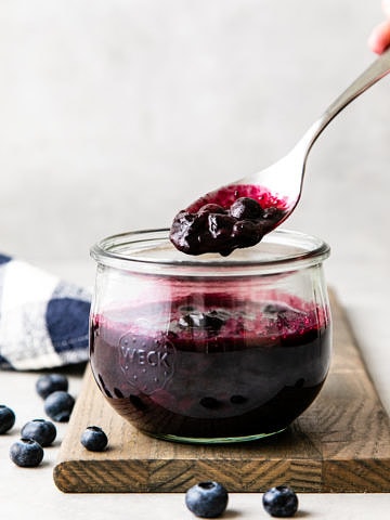 head on view of glass jar of blueberry compote with a spoon with scoop over top.