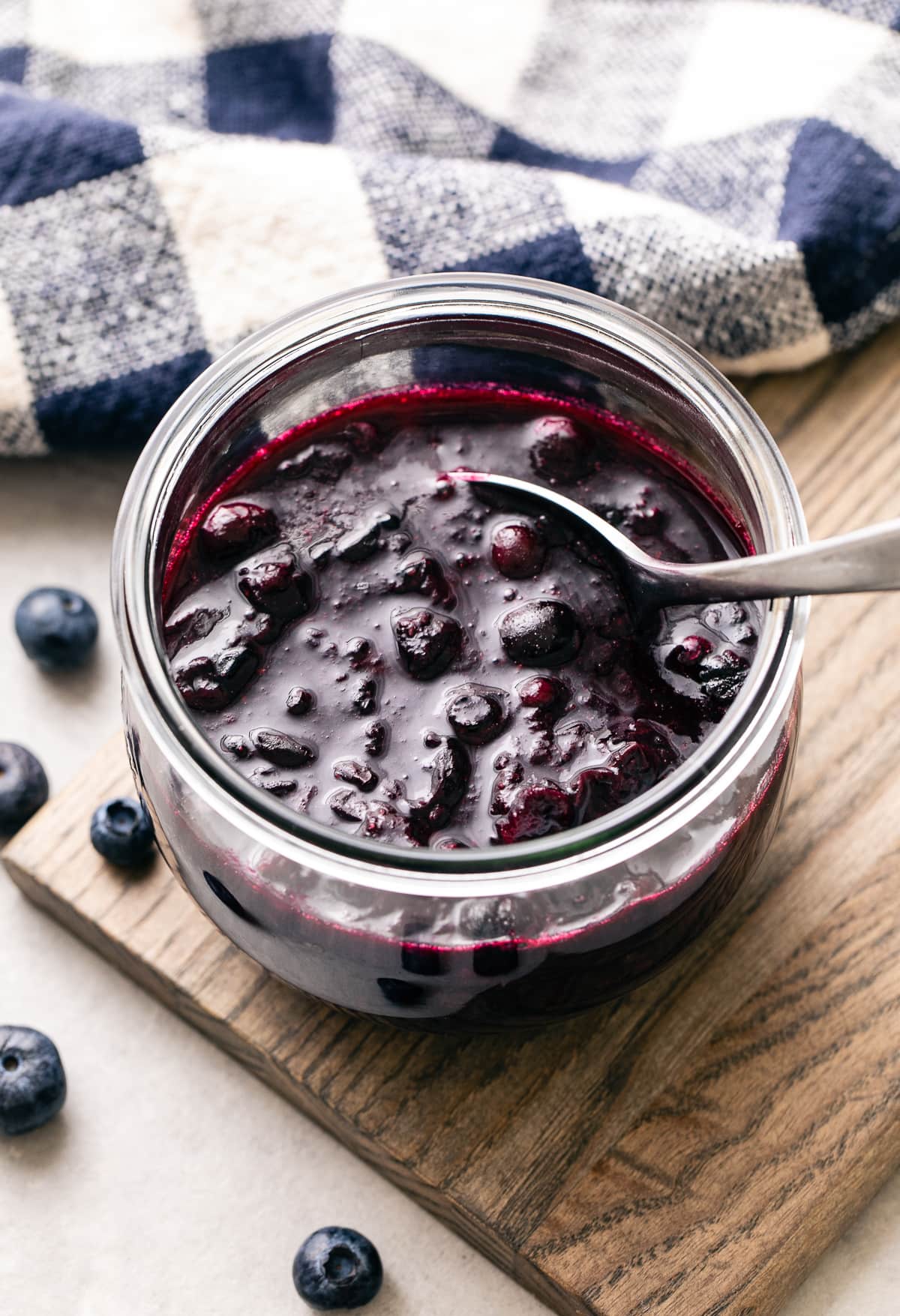 top down, side angle view of blueberry compote in a glass jar with spoon and items surrounding.