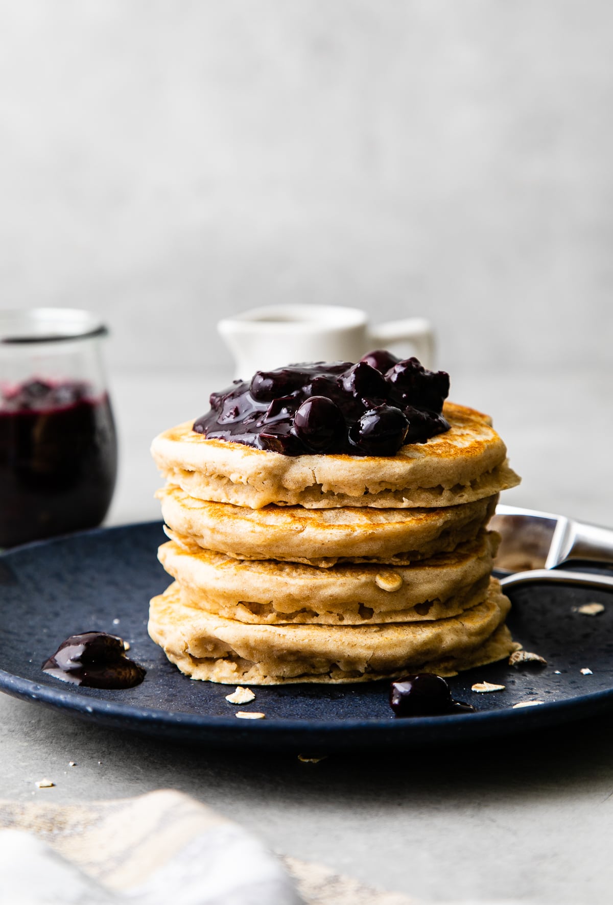 head on view stack of oatmeal pancakes topped with blueberry compote on a plate with items surrounding.
