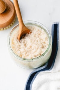 side angle view of sugar scrub in a jar with wooden spoon and items surrounding.