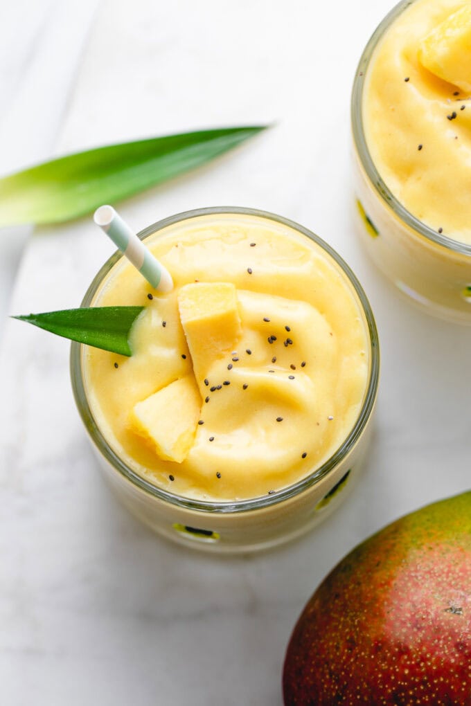 up close of pineapple banana mango smoothie in glass with straw and items surrounding.