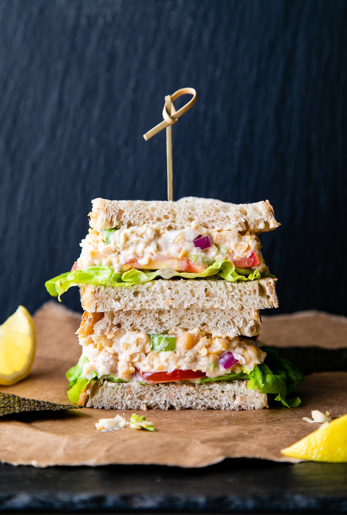head on view of chickpea 'tuna' salad sandwich cut in half and stacked with fresh lemon wedges.