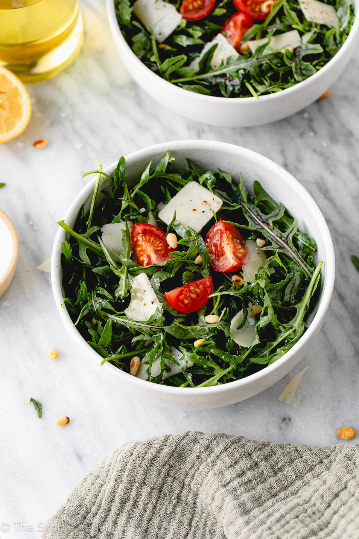 side angle view of arugula salad with cherry tomatoes, vegan cheese, and pine nuts in white bowls with items surrounding.