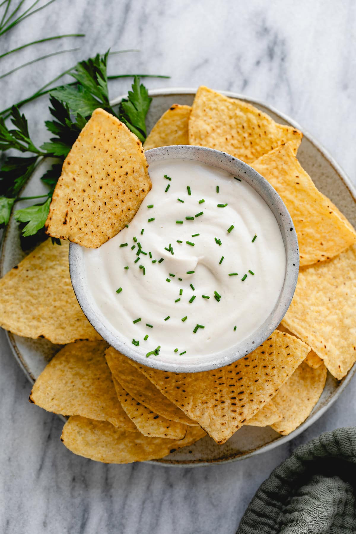 top down view of vegan sour cream in a bowl with tortilla chips and herbs surrounding.