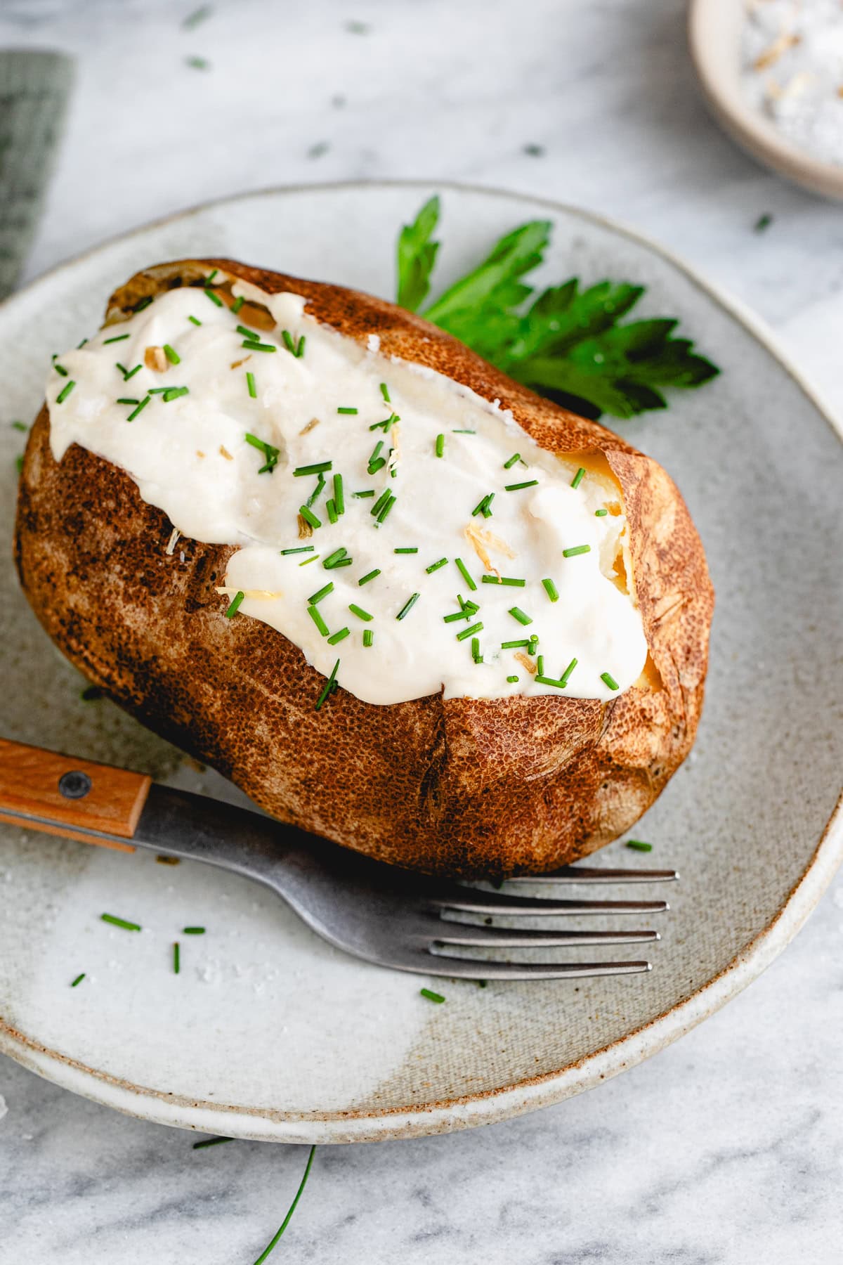 side angle view of plated baked potato with sour cream and chives with items surrounding.