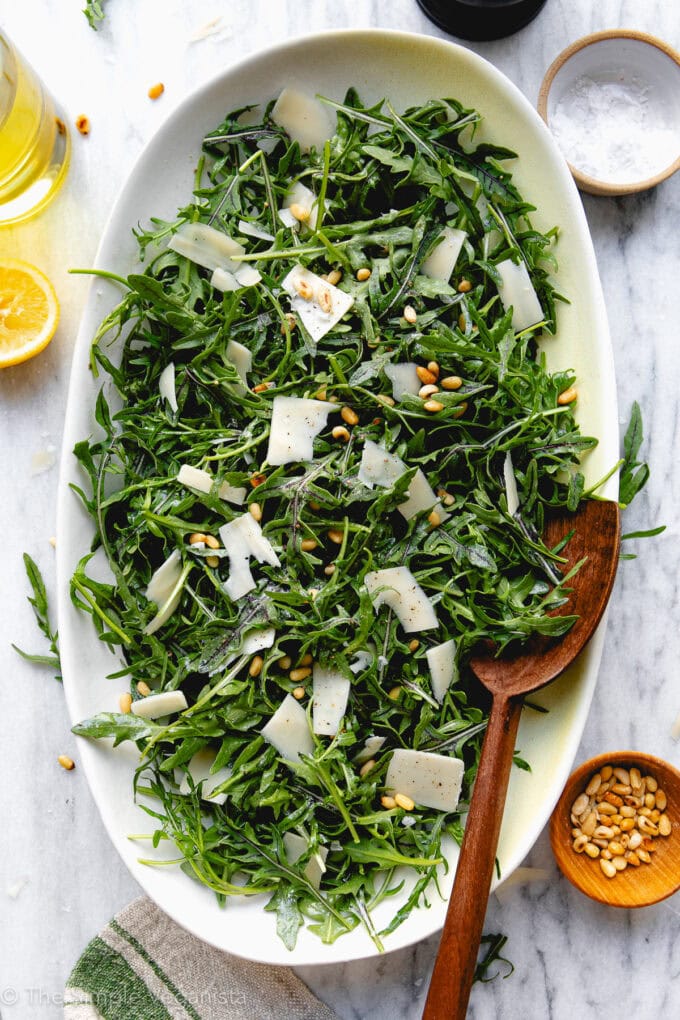 top down view of arugula salad with pine nuts and vegan Parmesan cheese on a white platter and items surrounding.