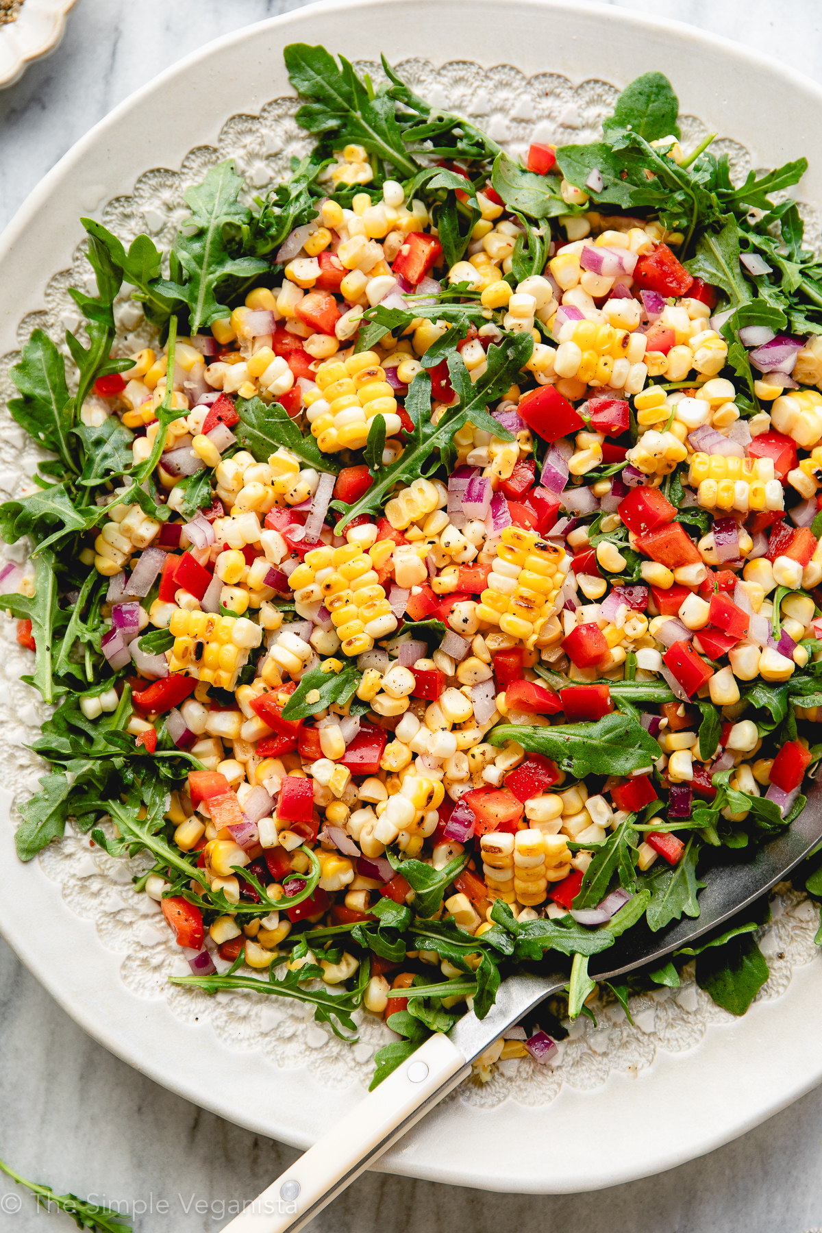 up close top down view of arugula corn salad on a white serving platter with large spoon and items surrounding.