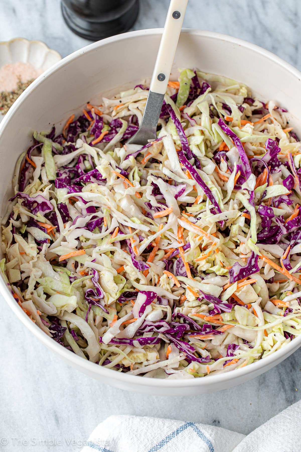 side angle view of vegan coleslaw in a large white bowl with spoon and items surrounding.