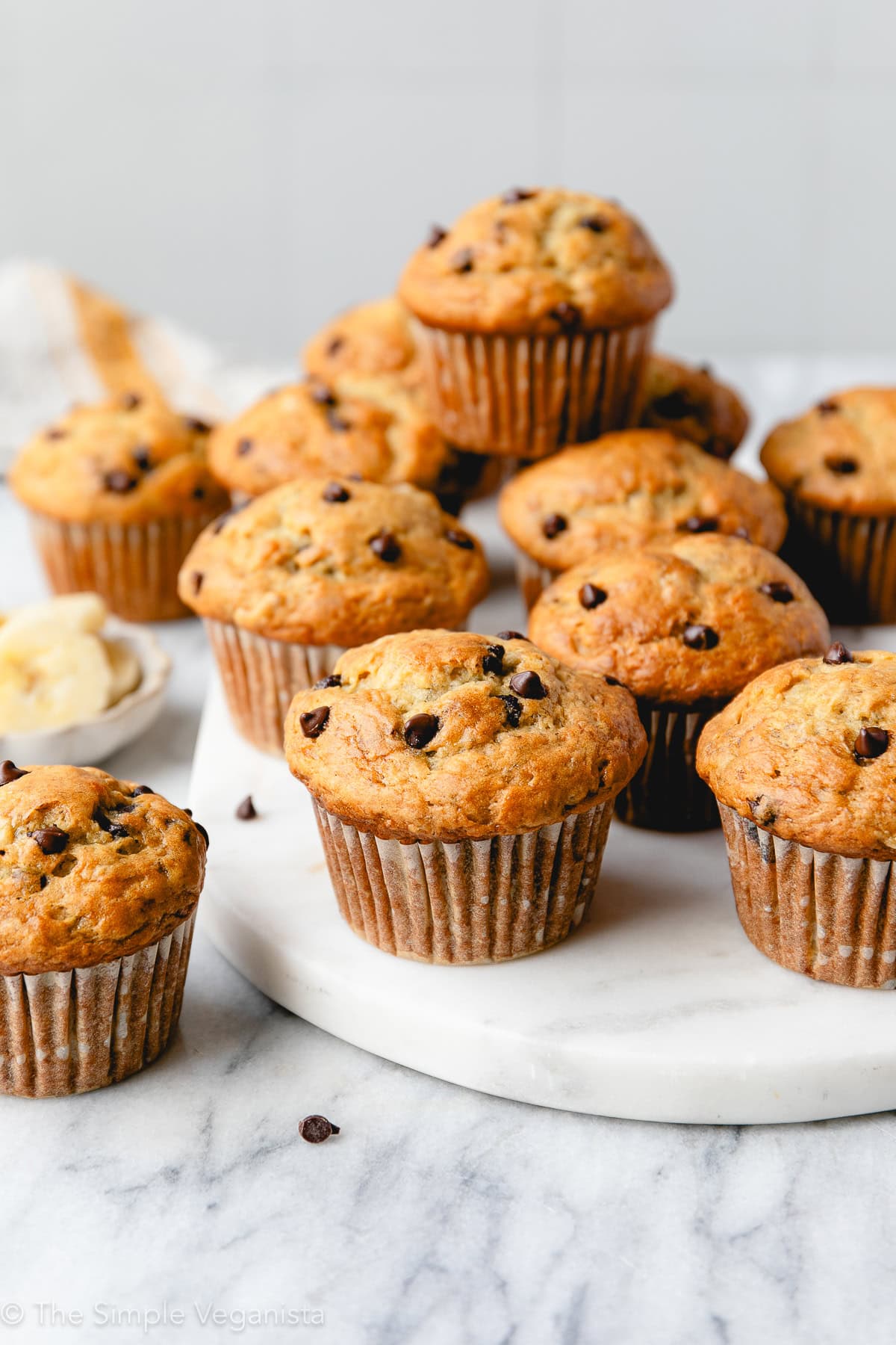 head on view of group of banana muffins with chocolate chips on a marble slab with other items surrounding.