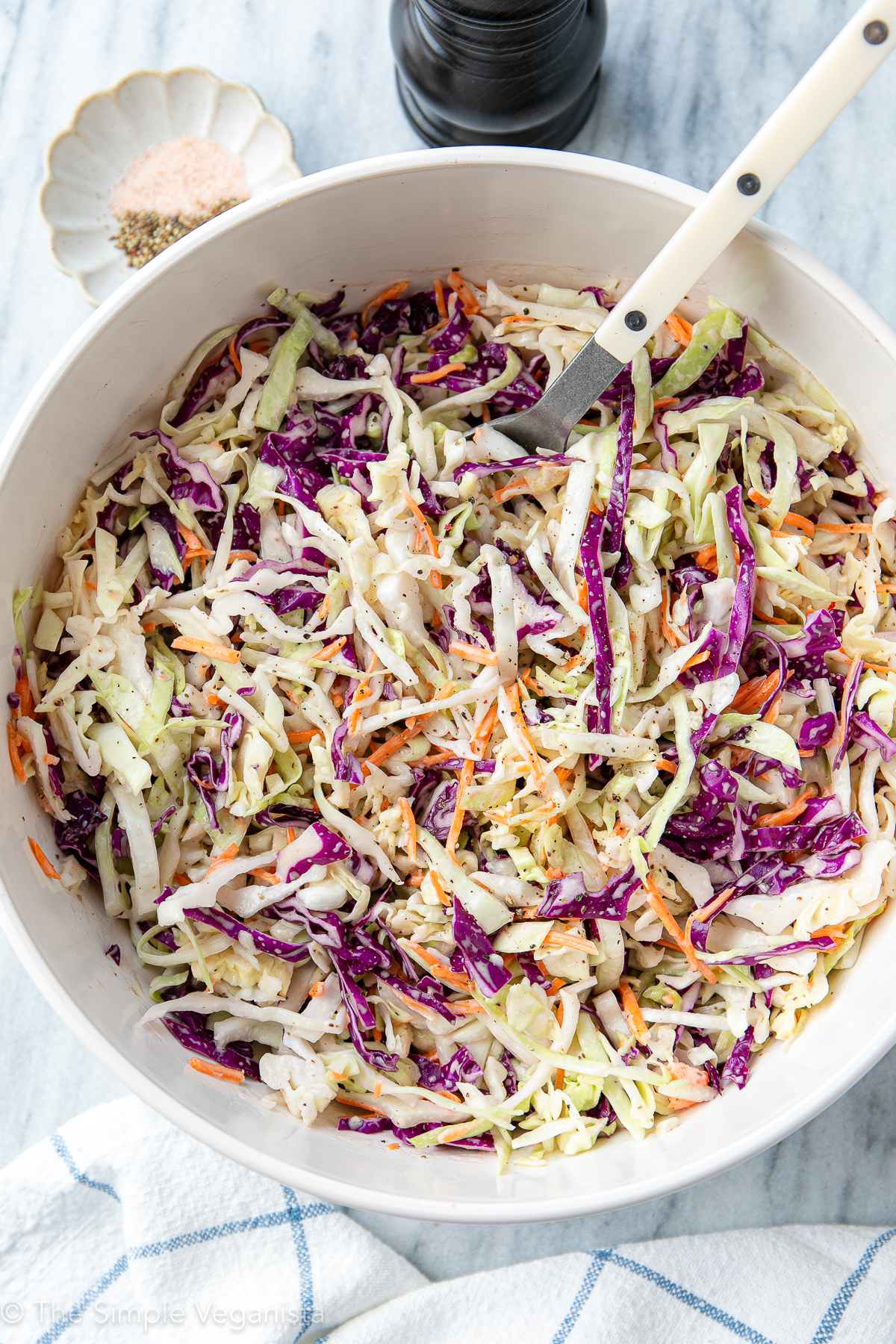 top down view of vegan coleslaw in a large white bowl with spoon and items surrounding.